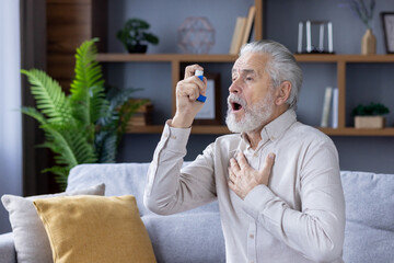 An elderly gray-haired man suffers from asthma. He sits at home on the couch and uses an inhaler