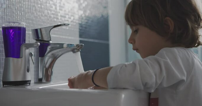 A young girl standing on a stool to reach the sink is diligently washing her hands with soap and water in a clean bathroom.