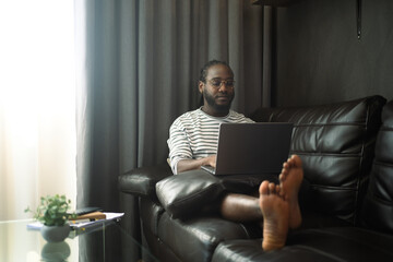 Focused African American male sitting on couch working remotely with laptop