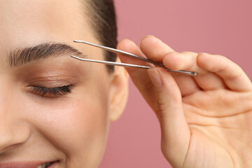 Eyebrow correction. Young woman with tweezers on pink background, closeup
