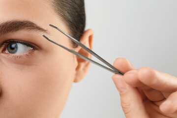 Eyebrow correction. Young woman with tweezers on light grey background, closeup