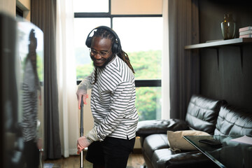 Happy African man listening to music in headphone while wiping floor with mop at home - 792669962