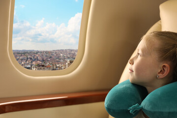 Cute little girl with travel pillow looking out of window in airplane during flight