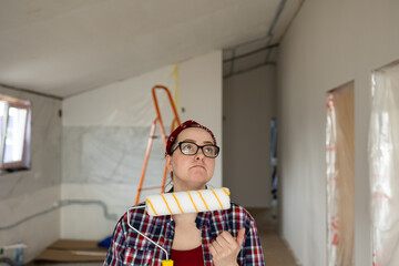 A woman in glasses with a paint roller in her hands looks skeptically up at the ceiling. High quality photo