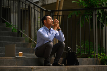 Thoughtful asian male entrepreneur sitting on stairs with bicycle and looking away