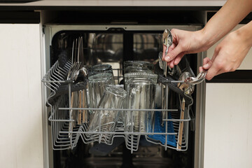 Woman loading dishwasher with glass and cutlery indoors, closeup