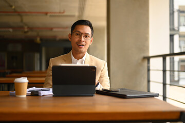 Portrait of Asian businessman in eyeglasses sitting with digital tablet and looking at camera - 792674925