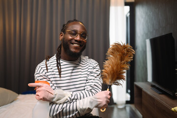 Happy smiling young African guy holding a feather duster ready for cleaning his apartment - 792676141