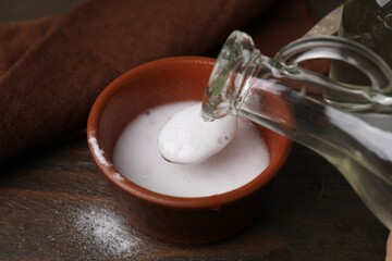 Pouring vinegar into spoon with baking soda over bowl at wooden table, closeup