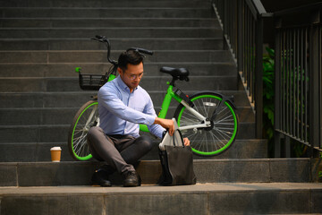 Shot of businessman with briefcase sitting on stairs in the city near his bicycle - 792680193