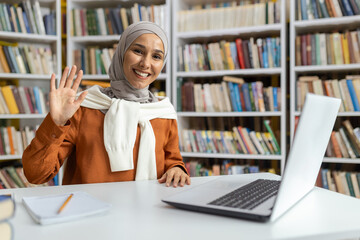 A cheerful Muslim woman in a hijab waves happily while participating in a video call at a library...