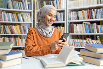 A cheerful Muslim woman smiling as she uses her smartphone amidst stacks of books in a bright, colorful library setting.