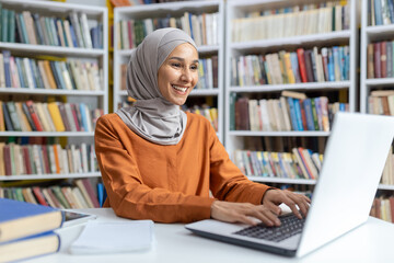 A cheerful Muslim woman wearing a hijab is studying in a library, using a laptop. Books in various...