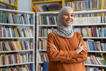 A cheerful Muslim woman in a hijab stands confidently with arms crossed in a vibrant library full of books, exuding a sense of intelligence and positivity. - obrazy, fototapety, plakaty