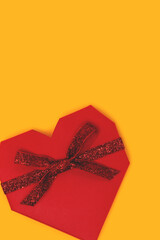 Red gift box in a heart shape with tied glittering ribbon on a yellow background.