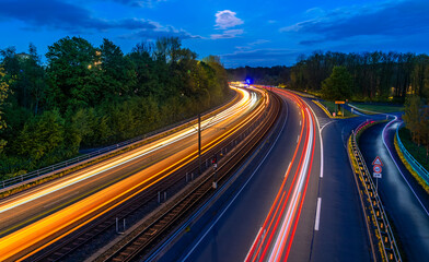 Blue hour panorama of “B54” expressway in the south of Dortmund in the Ruhr area (Germany). Curve with four lanes and double-track tram route in blue hour. Colorful light traces from train and cars.