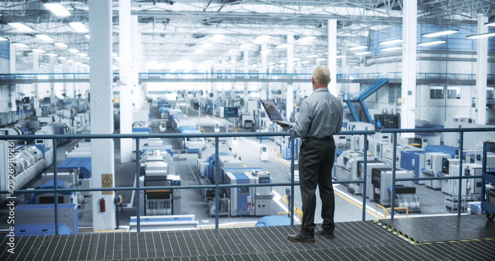 Leinwandbilder Experienced Male Engineer Standing on a Platform, Using Laptop Computer and Overlooking Production at a Modern Automated Electronics Manufacture with AI Support. Factory Executive at Work