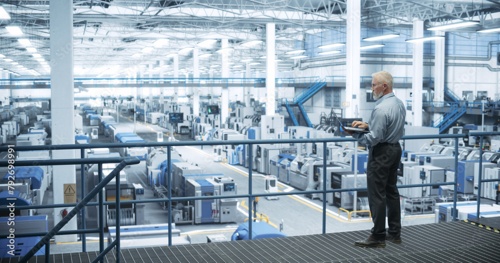 Poster experienced male engineer standing on a platform, using laptop computer and overlooking production a