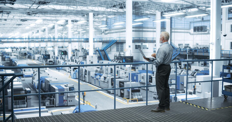 Naklejka premium Senior Engineer Standing on a Platform, Using Laptop Computer at an Electronics Factory. Machines are Undergoing Maintenance, Specialist Monitoring the Progress Through Online Software