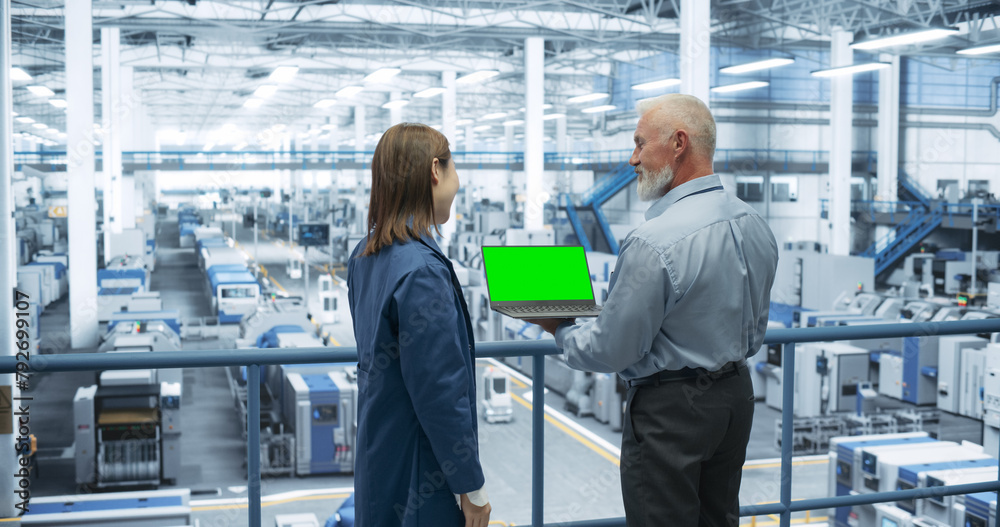 Fototapete Two Multiethnic Engineers Standing on a Platform, Using Laptop Computer with Green Screen at an Electronics Factory. Machines are Undergoing Maintenance, Specialists Monitoring the Progress