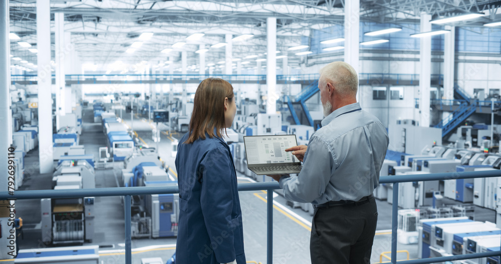 Aufkleber Two Multiethnic Engineers Standing on a Platform, Using Laptop Computer at an Electronics Factory. Machines are Undergoing Maintenance, Specialists Monitoring the Progress Through Online Software