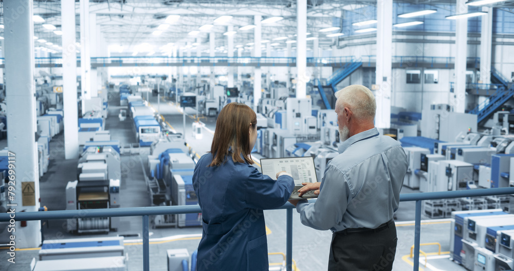 Poster Portrait of a Middle Aged Engineer and Happy Asian Scientist Using Laptop Computer and Talking in a Factory Facility with Equipment Producing Modern Electronic Components for Different Industries