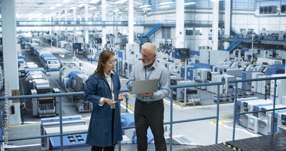 Aufkleber Two Multiethnic Engineers Standing on a Platform, Using Laptop Computer at an Electronics Factory. Machines are Undergoing Maintenance, Specialists Monitoring the Progress Through Online Software