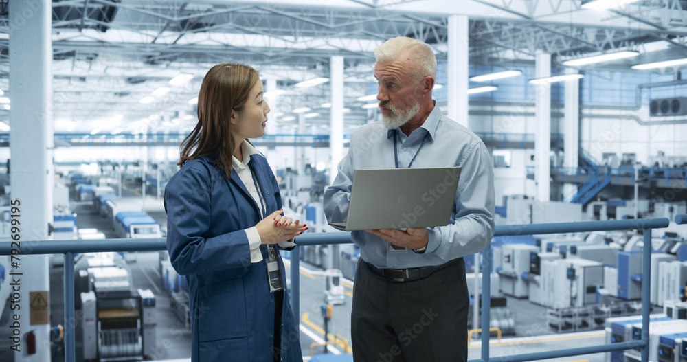 Poster Portrait of a Middle Aged Engineer and Happy Asian Scientist Using Laptop Computer and Talking in a Factory Facility with Equipment Producing Modern Electronic Components for Different Industries