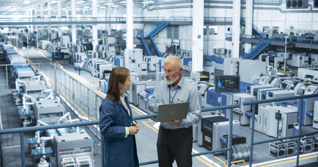 Two Multiethnic Engineers Standing on a Platform, Using Laptop Computer at an Electronics Factory. Machines are Undergoing Maintenance, Specialists Monitoring the Progress Through Online Software