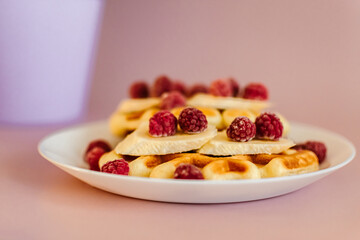 Viennese waffles with raspberries and bananas on a white plate next to yellow flowers in a pot. plain light background. homemade baking. recipe
