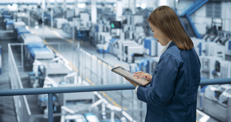 Fototapeta premium Experienced Female Engineer Standing on a Platform, Using Tablet Computer and Overlooking Production at a Modern Automated Electronics Manufacture with AI Support. Factory Manager at Work