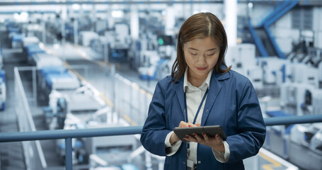 Young Asian Female Engineer Standing on a Platform, Using Tablet Computer at an Electronics Factory. Machines are Undergoing Maintenance, Specialist Monitoring the Progress Through Online Software
