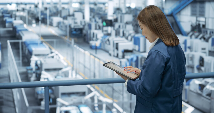 Experienced Female Engineer Standing on a Platform, Using Tablet Computer and Overlooking Production at a Modern Automated Electronics Manufacture with AI Support. Factory Manager at Work