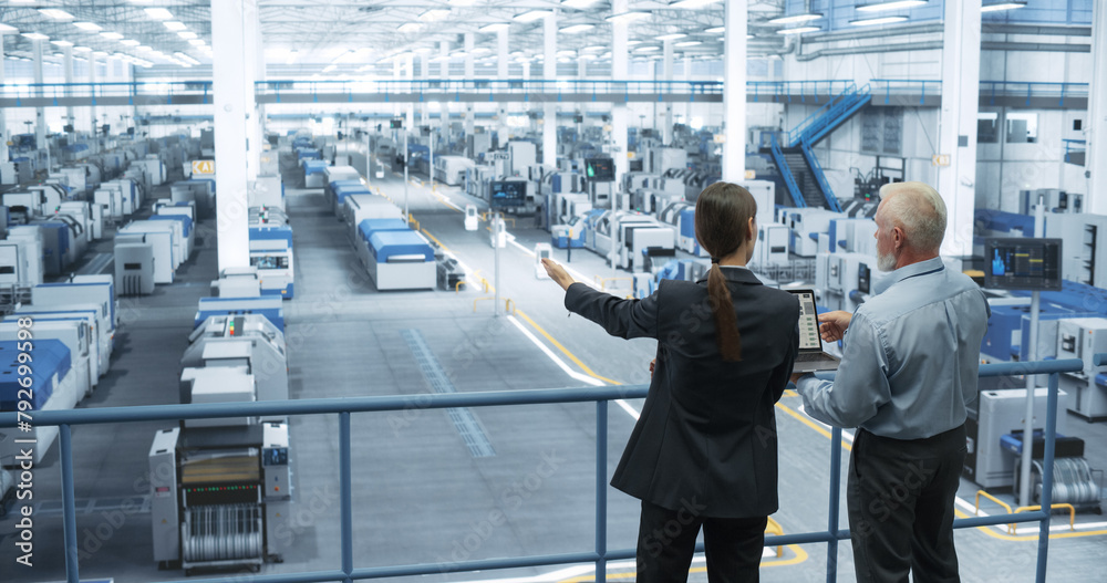 Aufkleber Middle Aged Engineer and Female Scientist Standing with Their Back, Using Laptop Computer and Talking in a Factory Facility with Equipment Producing Modern Technical Devices for Different Industries