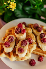 Viennese waffles with raspberries and bananas on a white plate next to yellow flowers in a pot. plain light background. homemade baking. recipe