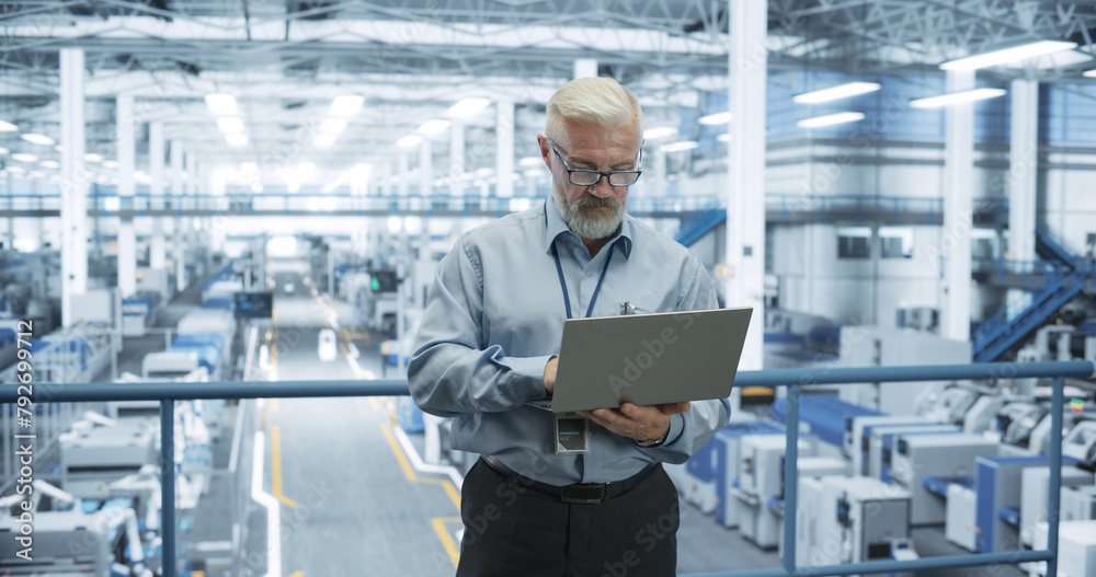 Fototapete Senior Engineer Standing on a Platform, Using Laptop Computer at an Electronics Factory. Machines are Undergoing Maintenance, Specialist Monitoring the Updates Through Online Software