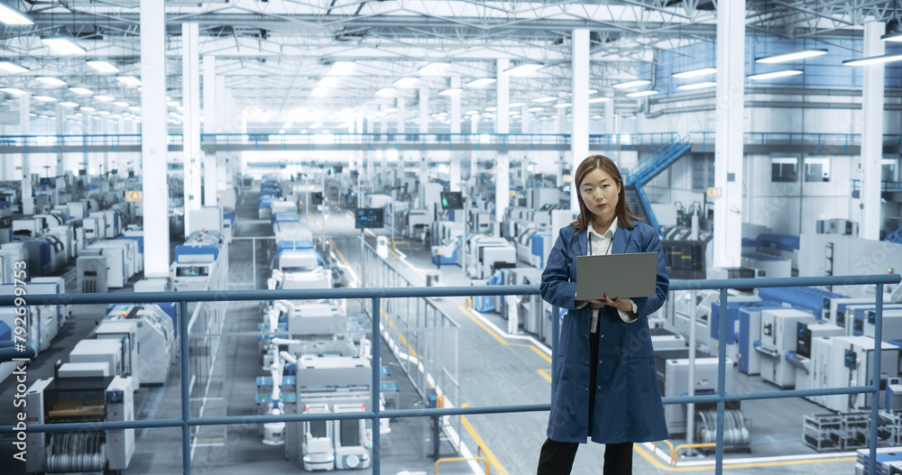 Aufkleber Shot in an Electronics Factory. Japanese Project Manager Working on Laptop at a Futuristic Industrial Manufacture with AI Robotic Arms. Specialist Monitoring Production on a Computer