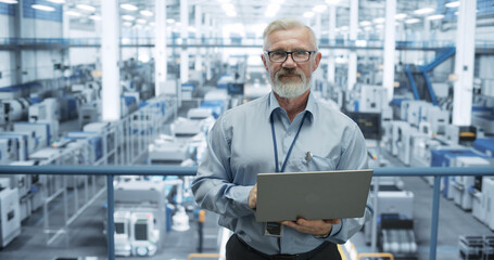 Portrait of a Senior Technician with a Beard and Glasses Using Laptop Computer and Looking at Camera. Industrial Male Specialist Wearing a Protective Vest, Working in a Modern Electronics Factory