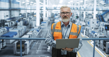 Portrait of a Senior Technician with a Beard and Glasses Using Laptop Computer and Looking at Camera. Industrial Male Specialist Wearing a Protective Vest, Working in a Modern Electronics Factory - 792699734