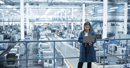 Experienced Female Engineer Standing on a Platform, Using Laptop to Monitor Data on a Computer, Check Reports and Plan a Research Strategy. Empowered Factory Manager at Work