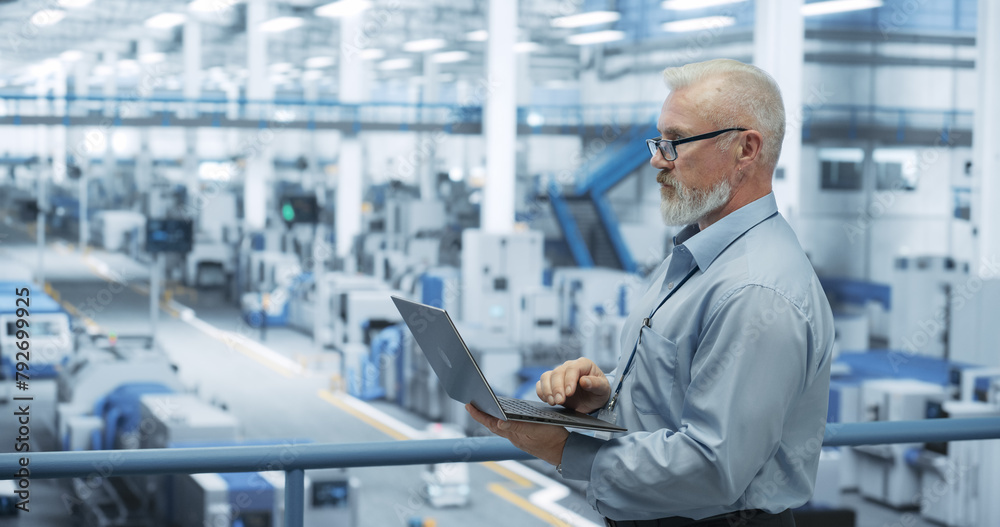 Aufkleber Portrait of a Handsome Middle Aged Engineer with Glasses Using Laptop Computer and Looking Around a Factory Facility with Equipment Producing Modern Electronic Components for Different Industries