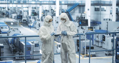 Two Female Technicians in Protective Coveralls Having a Conversation and Using Tablet Computer. Specialists Monitoring, Analyzing Conditions at a Modern Electronics Factory with Automated Robots