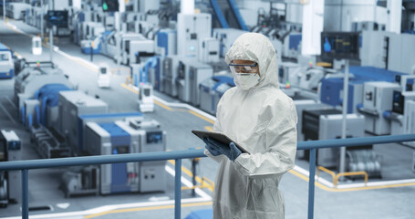 Female Technician in Protective Coveralls, Mask and Eyewear Using Tablet Computer at a Manufacturing Complex. Specialist Monitoring Conditions at a Modern Electronics Factory with Automated Robots - obrazy, fototapety, plakaty