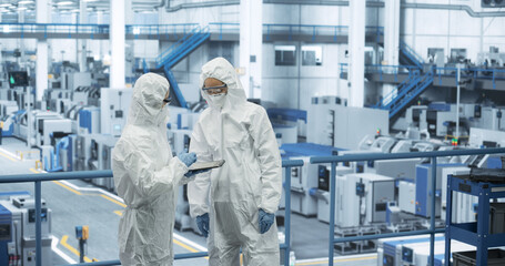 Two Female Technicians in Protective Coveralls Having a Conversation and Using Tablet Computer. Specialists Monitoring, Analyzing Conditions at a Modern Electronics Factory with Automated Robots - 792699978