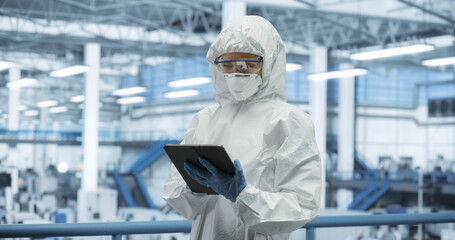 Female Technician in Protective Coveralls, Mask and Eyewear Using Tablet Computer at a Manufacturing Complex. Specialist Monitoring Conditions at a Modern Electronics Factory with Automated Robots