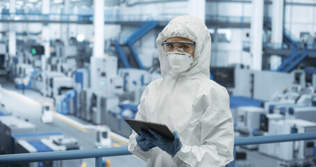 Female Worker Wearing Full-Body Protective Coveralls, Mask and Goggles at a Manufacturing Plant. Portrait of a Young Woman Specialists Using a Tablet Computer, Looking at Camera, Smiling - 792699996