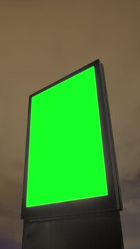 Timelapse, vertical video: blank vertical street green billboard poster or advertising pillar against fast moving warm clouds at night. Time lapse, green screen, copyspace and chroma key concept