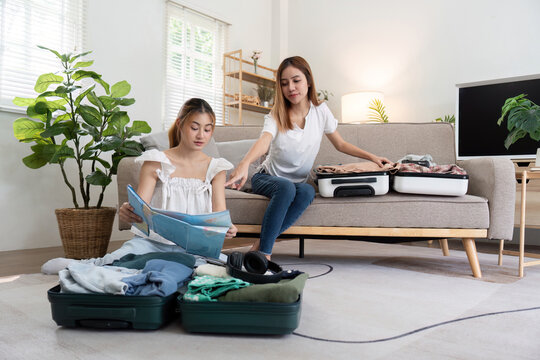 Young Asian woman packing clothes to the suitcase sitting on the sofa. Preparation for the summertime vacation. Two women are planning a trip and helping to prepare luggage to travel