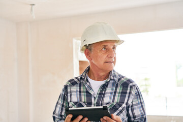 Senior male architect wearing hardhat holding tablet PC looking away inside incomplete house