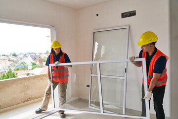 Full length of carpenters wearing safety workwear installing window frame in house construction site